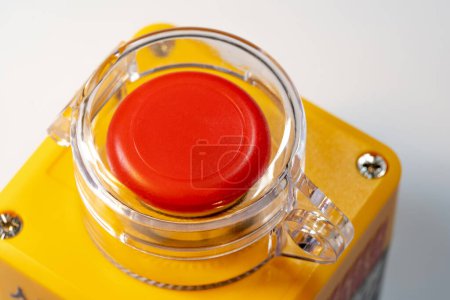 Detailed close-up of the upper part of a red emergency stop button with a transparent protective cover, mounted on a bright yellow industrial factory equipment machine control panel, workplace safety
