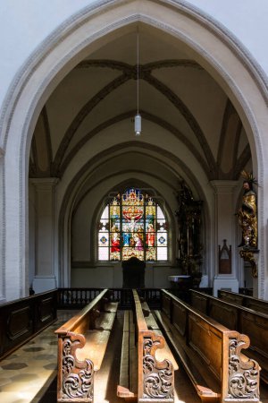 Téléchargez les photos : Détail des bancs de bois sculptés dans une église éclairée par la lumière du soleil avec un vitrail en arc coloré derrière et plafond voûté - en image libre de droit