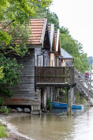 Téléchargez les photos : Petite barque amarrée dans l'eau d'un lac ou d'une rivière tranquille au-dessous de maisons en bois avec terrasse extérieure entourée de feuillage vert - en image libre de droit