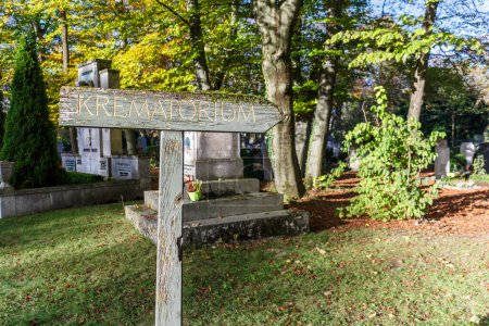 Téléchargez les photos : Krematorium Signe rustique en bois de langue allemande dans un cimetière pointant à droite avec des pierres tombales visibles derrière des arbres ombragés - en image libre de droit
