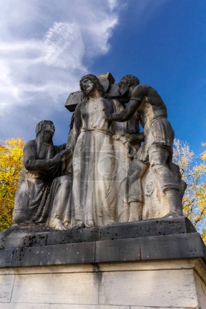 Téléchargez les photos : Statue en pierre de Jésus portant la croix à sa crucifixion vue à angle bas avec des feuilles d'automne colorées et un ciel bleu - en image libre de droit
