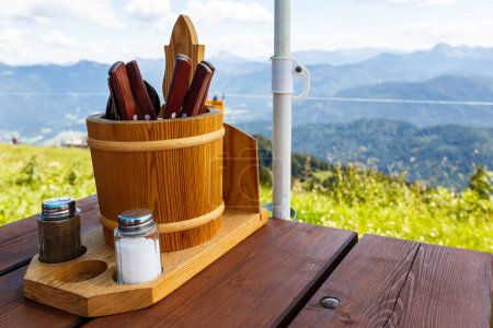 Photo for Cutlery and condiments in a rustic wooden container on a restaurant table overlooking a scenic landscape of mountains and valleys - Royalty Free Image