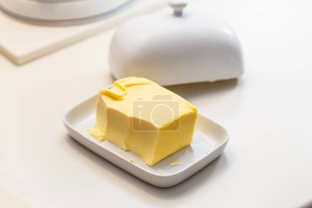 Téléchargez les photos : Pat de beurre frais dans un plat de beurre en céramique avec le couvercle retiré sur une table blanche - en image libre de droit