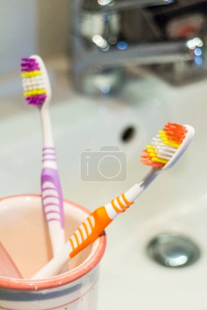 Téléchargez les photos : Deux brosses à dents en plastique dans une tasse sur le côté d'un lavabo blanc dans une salle de bain avec dof peu profond - en image libre de droit