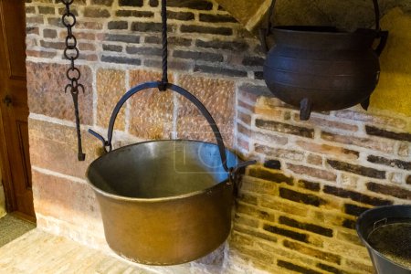 Téléchargez les photos : Divers pots de cuisson en métal suspendus dans un foyer avec un chaudron noir et un pot en laiton suspendu aux chaînes et aux crochets - en image libre de droit