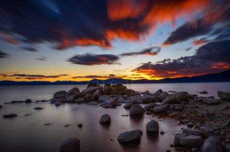 Sunset above rocky beach of Lake Tahoe in California with Sierra Nevada Mountains in the background. Long exposure.
