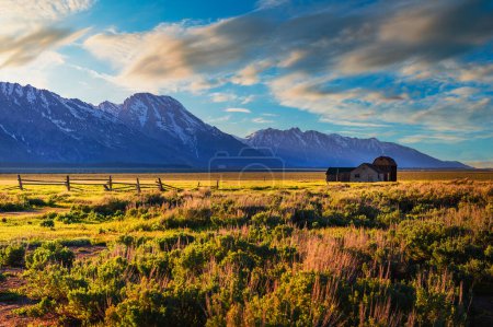 Téléchargez les photos : Coucher de soleil estival sur une ferme historique et la grange Thomas Murphy à Mormon Row dans le parc national de Grand Teton, avec la chaîne de montagnes de Teton enneigée en arrière-plan. - en image libre de droit
