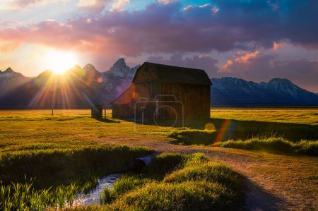 Téléchargez les photos : Coucher de soleil estival sur la grange historique Thomas Murphy à Mormon Row dans le parc national Grand Teton, avec une chaîne de montagnes Teton enneigée en arrière-plan et un cours d'eau au premier plan. - en image libre de droit