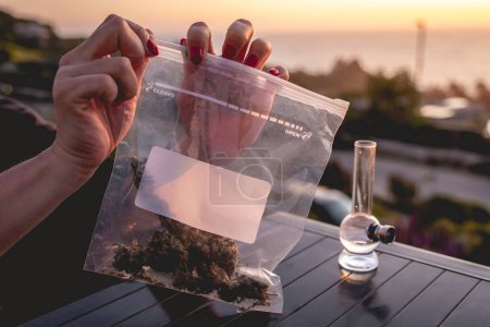 Téléchargez les photos : Mains avec des ongles rouges tenant un sac en plastique avec de grands bourgeons de marijuana (et de l'espace de copie), bong (tuyau d'eau de mauvaise herbe) et beau coucher de soleil ciel et mer - en image libre de droit