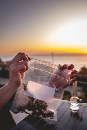 Téléchargez les photos : Mains avec des ongles rouges tenant un sac en plastique avec de grands bourgeons de marijuana (et de l'espace de copie), bong (tuyau d'eau de mauvaise herbe) et beau coucher de soleil ciel et mer - en image libre de droit