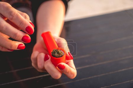 Téléchargez les photos : Mains avec des ongles rouges mettant un bourgeon de mauvaises herbes dans un tuyau en plastique orange marijuana et un briquet orange avec une belle lumière du coucher du soleil - en image libre de droit