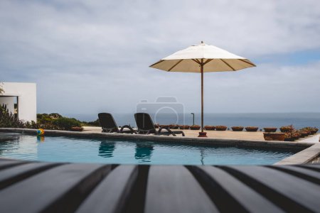 Téléchargez les photos : Chaise de plage devant une grande piscine en terrasse avec plus de chaises longues, parasol et l'océan dans une soirée d'été - en image libre de droit