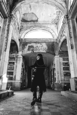 Téléchargez les photos : Femme gothique foncé avec robe noire dans un grand mausolée abandonné de style néoclassique avec niches, arches et lumière du soleil (en noir et blanc) - en image libre de droit