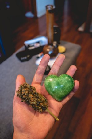 Téléchargez les photos : Main tenant une pierre de jade verte en forme de cœur et un gros bourgeon de marijuana (tête de mauvaise herbe) avec fond avec des outils de fumage de mauvaises herbes - en image libre de droit