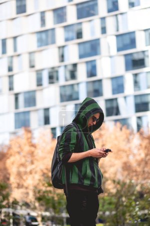 Téléchargez les photos : Jeune homme sérieux avec sweat à capuche vert, lunettes de soleil rondes et sac à dos bavardant dans un téléphone mobile dans un parc - en image libre de droit