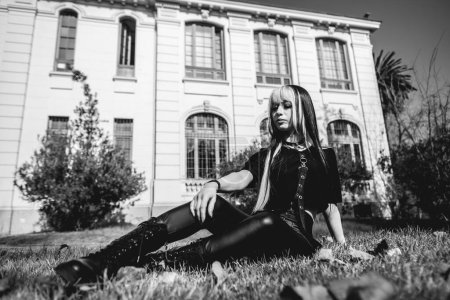 Téléchargez les photos : Portrait d'un modèle jeune et maigre avec de longs cheveux blonds et noirs, des vêtements noirs et des bottes noires assises dans le parc dans la journée avec bâtiment vieux siècles - en image libre de droit