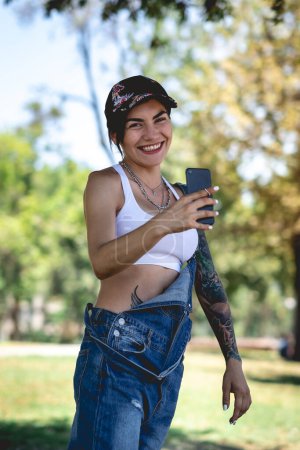 Photo for Happy, sexy and tattooed young girl with cap, denim dungarees and white crop top smiling, laughing and holding a phone in the park in a sunny day - Royalty Free Image
