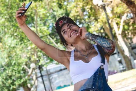 Photo for Happy, sexy and tattooed young girl with cap, denim dungarees and white crop top smiling, laughing and making a video call on the phone in the park in a sunny day - Royalty Free Image