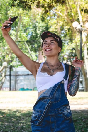 Photo for Happy, sexy and tattooed young girl with cap, denim dungarees and white crop top smiling, laughing and making a video call on the phone in the park in a sunny day - Royalty Free Image