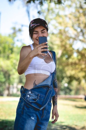 Photo for Happy, sexy and tattooed young girl with cap, denim dungarees and white crop top smiling, laughing and holding a phone in the park in a sunny day - Royalty Free Image