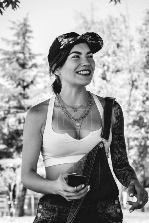 Photo for Happy, sexy and tattooed young girl with cap, denim dungarees and white crop top smiling, laughing and holding a phone in the park in a sunny day (in black and white) - Royalty Free Image