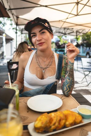 Photo for Happy, sexy and tattooed young girl with cap, denim dungarees and white crop top smiling in a restaurant table with fried pumpkin in panko - Royalty Free Image
