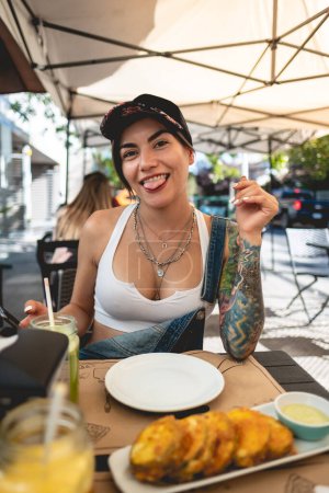 Photo for Happy, sexy and tattooed young girl with cap, denim dungarees and white crop top smiling in a restaurant table with fried pumpkin in panko - Royalty Free Image