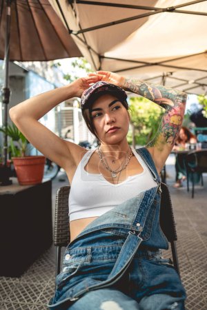 Photo for Serious, sexy and tattooed young girl with cap, denim dungarees and white crop top seated in a restaurant table - Royalty Free Image