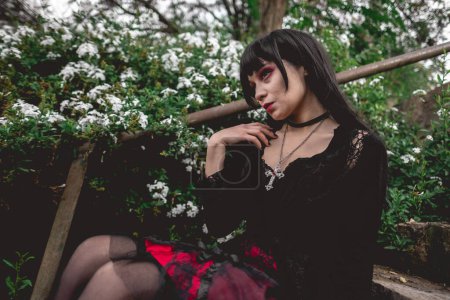 Téléchargez les photos : Jeune et maigre fille hispanique gothique avec robe noire et jupe rouge assis dans un escalier en pierre à côté de fleurs blanches dans la forêt - en image libre de droit