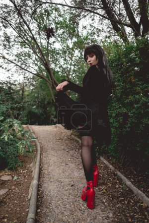 Téléchargez les photos : Jeune et maigre fille hispanique gothique avec robe noire, collants et chaussures plates-formes rouges marchant dans un chemin dans la forêt - en image libre de droit