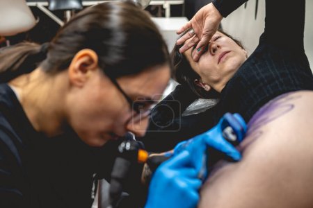 Téléchargez les photos : Jeune tatoueuse concentrée avec des lunettes faisant un tatouage, avec une machine à tatouer, d'un grand tigre, dans la cuisse et les hanches d'une belle femme souffrant de douleurs persistantes - en image libre de droit
