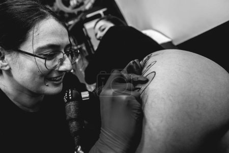 Téléchargez les photos : Jeune artiste tatoueuse souriante avec des lunettes faisant un tatouage, avec machine à tatouer, d'un grand tigre, dans la cuisse et les hanches d'une belle femme (en noir et blanc) - en image libre de droit