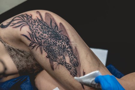 Foto de Joven artista del tatuaje manos con guantes azules limpieza de un tatuaje de un tigre grande, en el muslo y las caderas de una hermosa mujer - Imagen libre de derechos