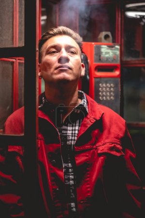 Téléchargez les photos : Portrait d'un jeune et séduisant mannequin veste rouge fumant une cigarette dans une cabine téléphonique rouge dans la ville dans la nuit - en image libre de droit