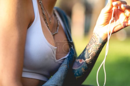Photo for Details of a rude, sexy and tattooed young girl with denim dungarees and white crop top seated in the park making a call with headphones in a sunny day - Royalty Free Image