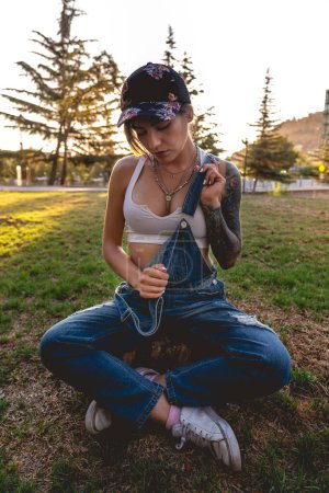 Photo for Rude, sexy and tattooed young girl with cap, denim dungarees, white crop top and white sneakers seated in a trunk in the park in a sunny day - Royalty Free Image