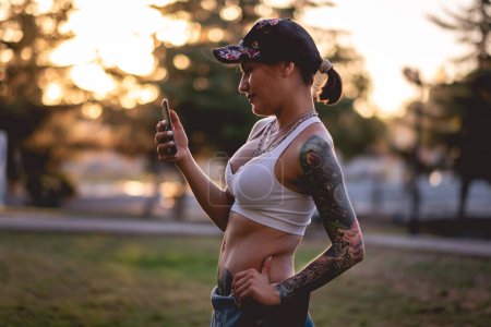 Photo for Rude, sexy and tattooed young girl with cap, denim dungarees and white crop top smiling and making a video call on the phone in the park in the sunset - Royalty Free Image