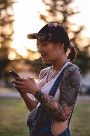 Photo for Rude, sexy and tattooed young girl with cap, denim dungarees and white crop top smiling and chatting on the phone in the park in the sunset - Royalty Free Image