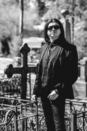Téléchargez les photos : Stylish dark gothic guy with black clothes in front of a grave in cemetery in a sunny day (in black and white) - en image libre de droit