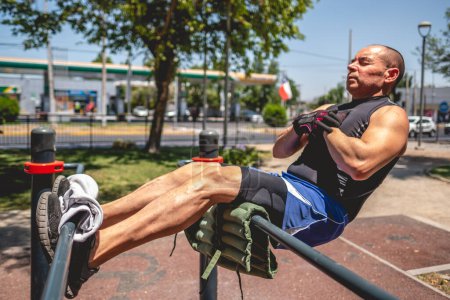 Téléchargez les photos : Muscular, healthy and fit elderly man training in parallel bars in a street workout park with trees and blue sky in a sunny day - en image libre de droit
