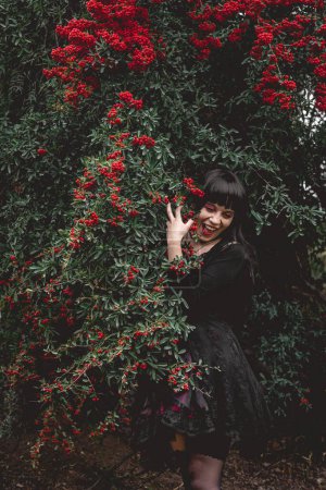Téléchargez les photos : Young and skinny white goth girl with long black hair and black dress happy and laughing between big rowan berries trees in the forest - en image libre de droit