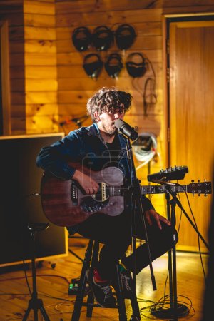 Téléchargez les photos : Young rocker musician with messy hair playing a wood electro acoustic guitar and singing in a music studio - en image libre de droit