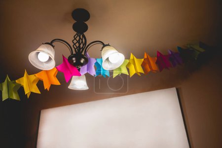 Téléchargez les photos : Lampe sur le toit avec trois décorations claires et colorées d'étoiles en papier pour une célébration et toile blanche vide dans le mur avec espace de copie - en image libre de droit