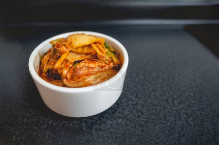 Téléchargez les photos : Kimchi de laitue traditionnelle épicée dans un bol blanc dans une table dans la cuisine avec un grand espace de copie (kimchi : légumes marinés et fermentés, ail, sel, gingembre, piments, etc.) - en image libre de droit