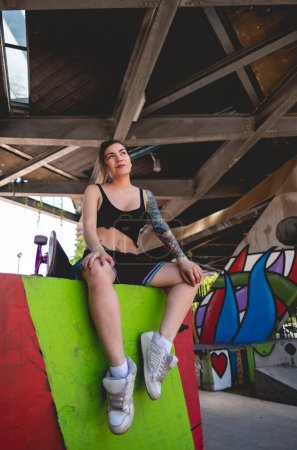 Téléchargez les photos : Style rebelle : jeune femme sexy avec haut noir et court, tatouages et skateboard en skate park urbain avec graffitis - en image libre de droit