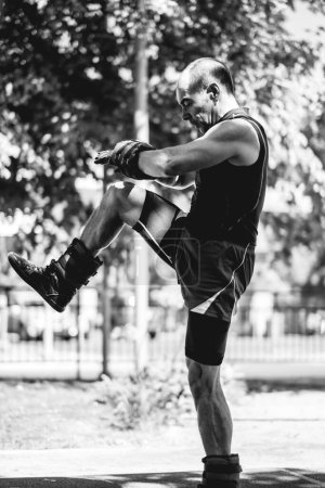 Téléchargez les photos : Sain et actif : homme âgé sans âge et musclé avec des poids corporels de sable entraînement dans le parc d'entraînement de rue le matin (en noir et blanc) - en image libre de droit