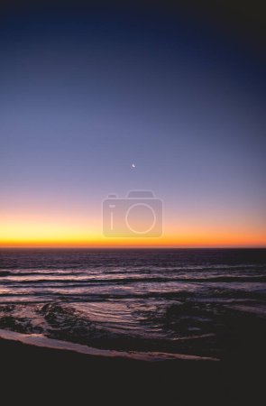 Téléchargez les photos : Tranquillité océanique : scène du croissant de lune blanc illuminant l'océan violet foncé, tandis que les vagues s'écrasent doucement sur le sable de la plage, sous un ciel serein de coucher de soleil - en image libre de droit