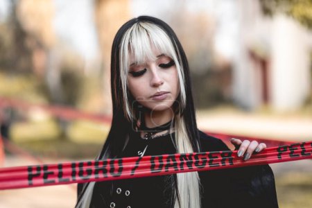 Téléchargez les photos : Dangereusement élégant : jeune femme en accessoires gothique roches tenue noire et pose derrière une ligne de danger rouge dans le parc - en image libre de droit