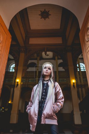 Téléchargez les photos : Jeune femme aux cheveux noirs et blonds et veste rose à l'entrée d'un musée public et intérieur vieux siècles à l'architecture néoclassique - en image libre de droit