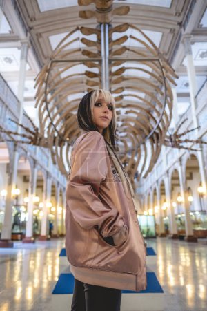 Téléchargez les photos : Jeune femme aux cheveux noirs et blonds et veste rose dans un ancien musée public intérieur à l'architecture néoclassique et un grand squelette de baleine - en image libre de droit